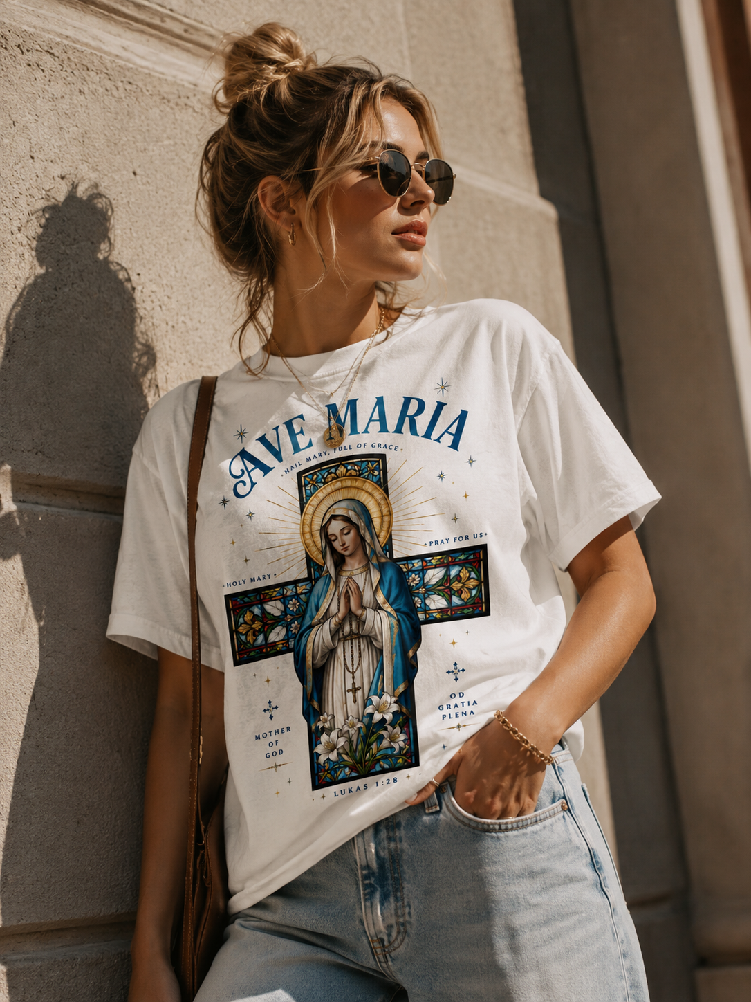 A woman wearing a white graphic t-shirt with a colorful 'Ave Maria' religious print, paired with light-wash jeans and sunglasses, leaning against a stone wall.
