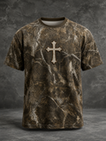 A short-sleeve crewneck t-shirt featuring a brown and beige distressed camouflage pattern with a raised metallic-look cross graphic on the chest, displayed against a dark gray textured studio background.