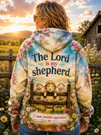 A woman stands outdoors at sunset wearing a hooded sweatshirt from the back. The hoodie features a colorful pastoral print with flowers, sheep in a fence, and the text 'The Lord is my shepherd.