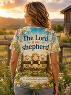 A woman stands outdoors in a field at sunset wearing a graphic T-shirt. The back of the shirt features a printed scene of sheep behind a wooden fence with the text 'The Lord is my shepherd' and smaller text below.