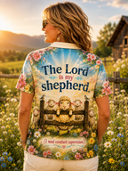 A woman stands outdoors in a sunlit field wearing a short-sleeve collared shirt from the back. The shirt has a colorful pastoral print with sheep, flowers, birds, and the text 'The Lord is my shepherd' and '(I need constant supervision)'.
