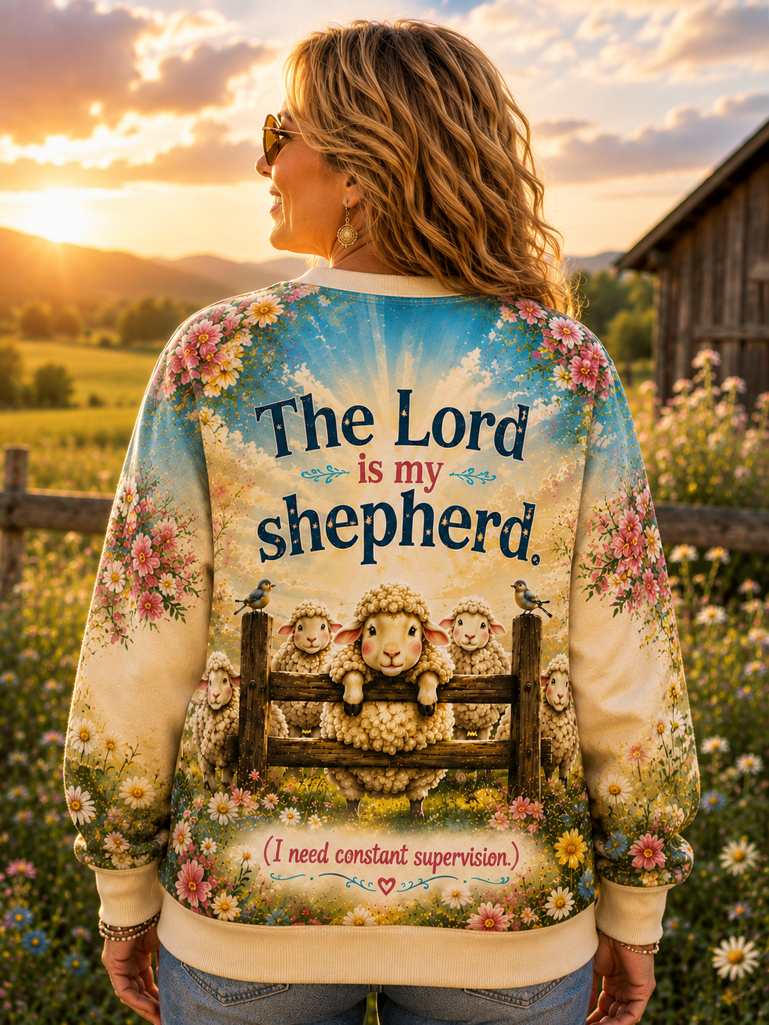A woman stands outdoors in a field at sunset wearing a cream-colored sweatshirt with a detailed graphic on the back. The graphic features the text 'The Lord is my shepherd' above an illustration of a sheep on a fence with other sheep, surrounded by floral designs.
