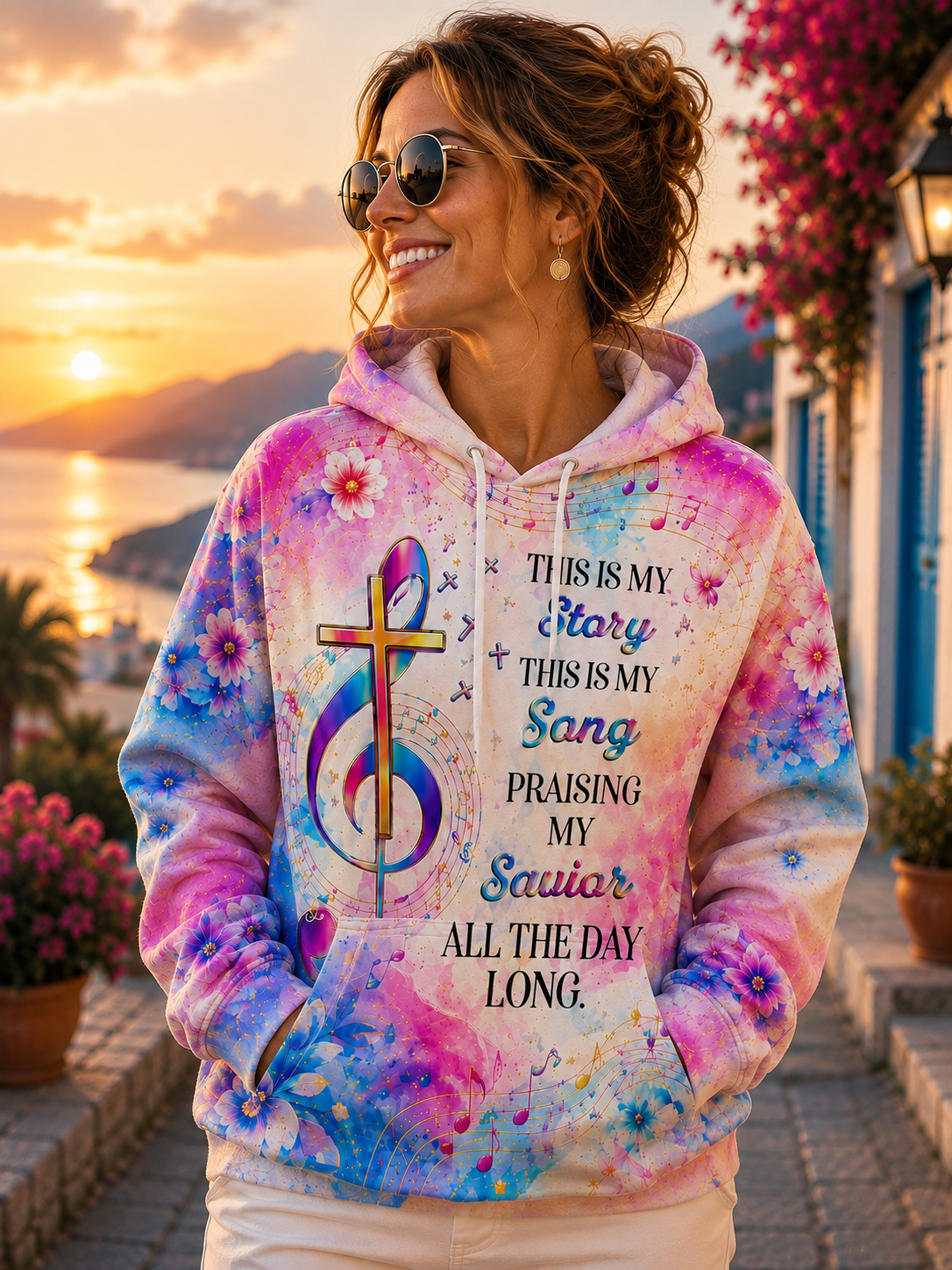 A woman wearing a colorful tie-dye hoodie with a cross, musical notes, and text that reads 'THIS IS MY Story THIS IS MY Sang Praising My Saiyar ALL THE DAY LONG.' She is standing outdoors on a cobblestone street at sunset with flowers and a coastal background.
