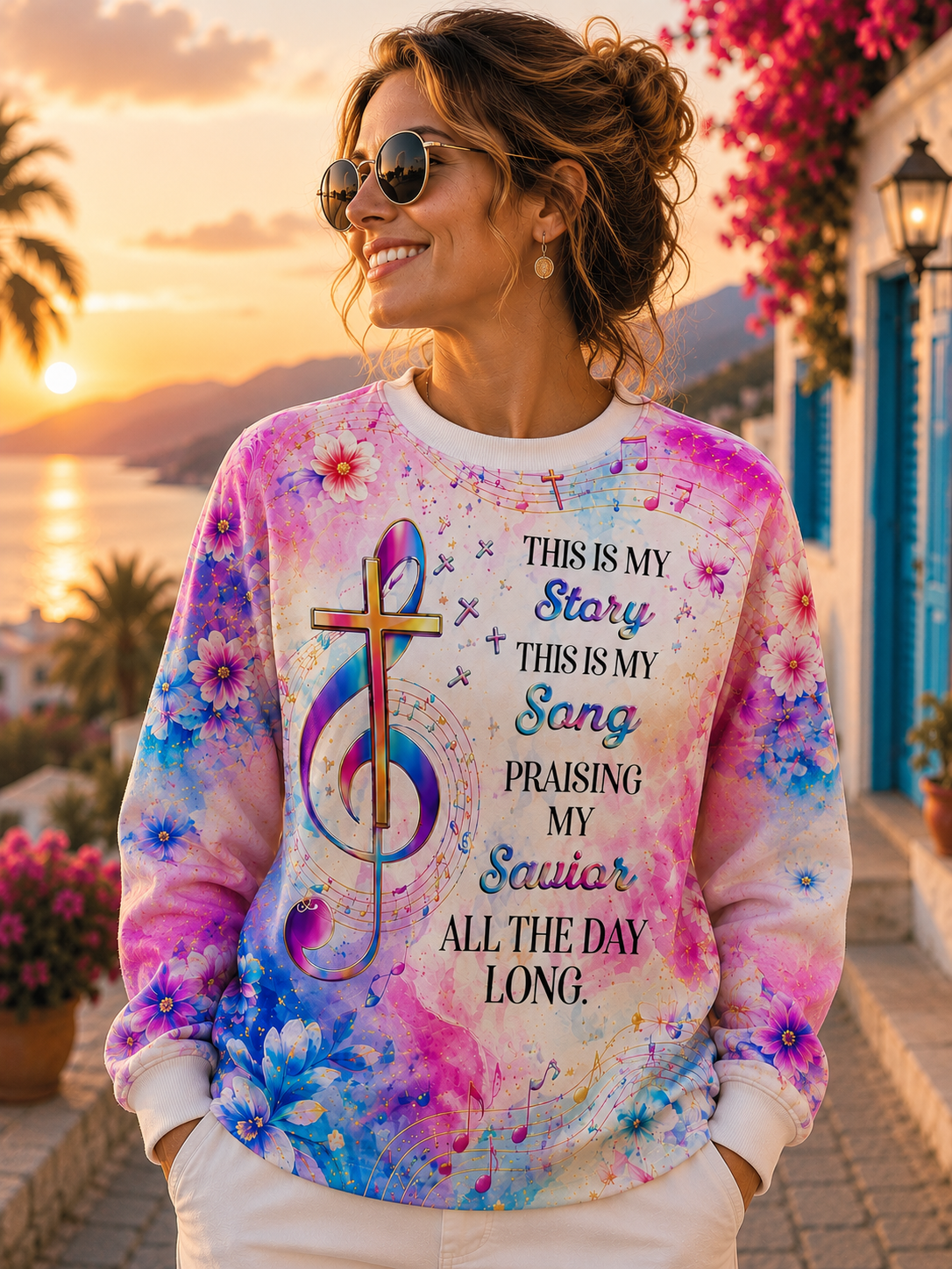 A woman wearing a colorful floral graphic sweatshirt with a cross and musical note design, standing outdoors in front of a sunset by the sea.