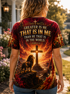 A woman viewed from behind wearing a short-sleeve graphic T-shirt with a fiery red and orange design. The shirt features a large cross, a lion, and the text 'GREATER IS HE THAT IS IN ME THAN HE THAT IS IN THE WORLD.